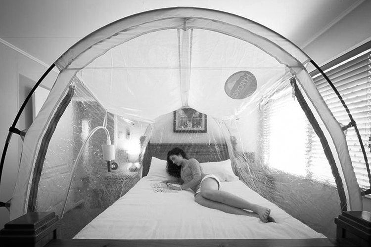 A woman lies comfortably on a bed, reading a book inside a transparent, dome-shaped altitude training tent.