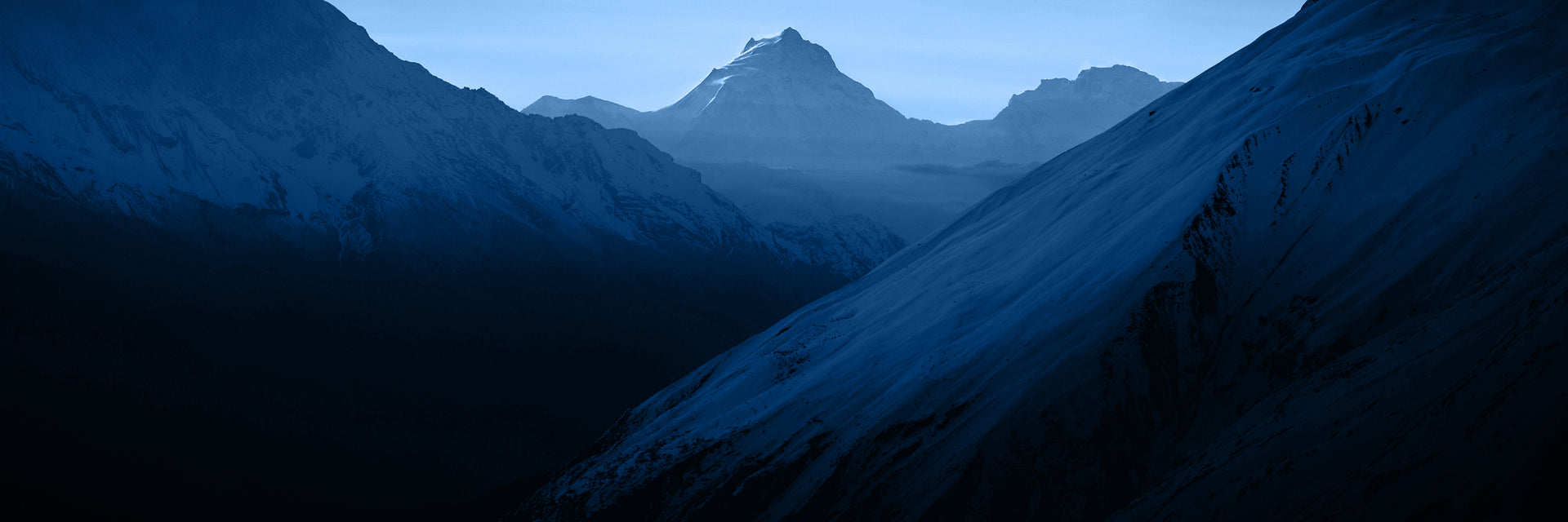 A wide, majestic view of dark, snow-dusted mountain ranges at dusk or dawn, with a prominent, jagged peak silhouetted against a lighter sky in the center.