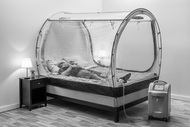A full shot of a bedroom with a transparent altitude training tent set up over a bed, with a person lying inside and an air generator unit beside it.