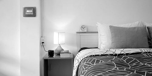 A modern bedroom scene in black and white, featuring a bed with a patterned duvet, a nightstand with a lamp and alarm clock, and a wall-mounted display.